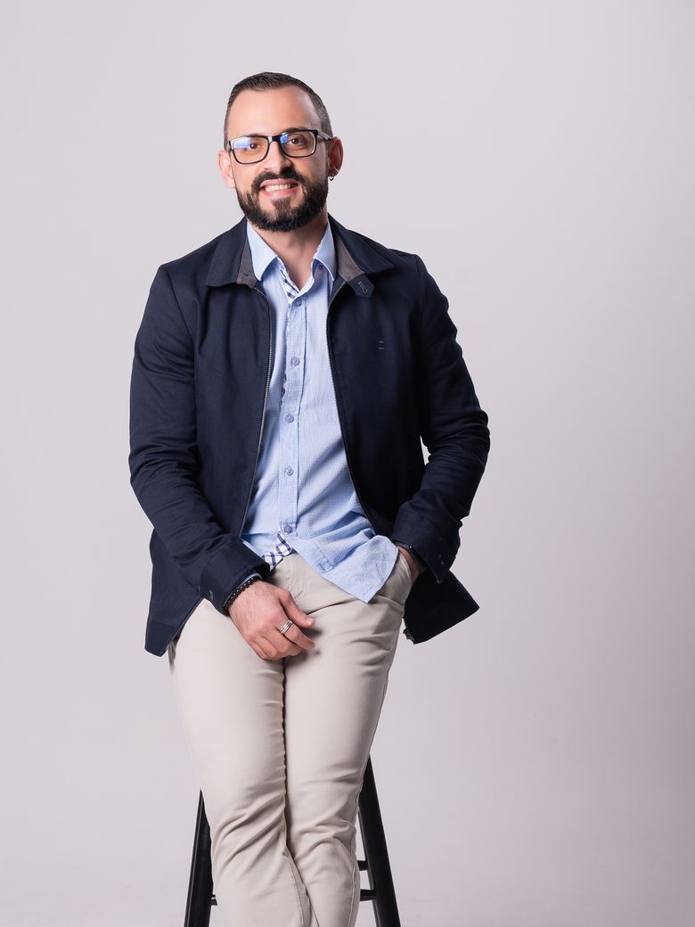 Caio Victor sentado num banco alto em estúdio claro, de jaqueta azul-marinho e camisa azul, sorrindo.