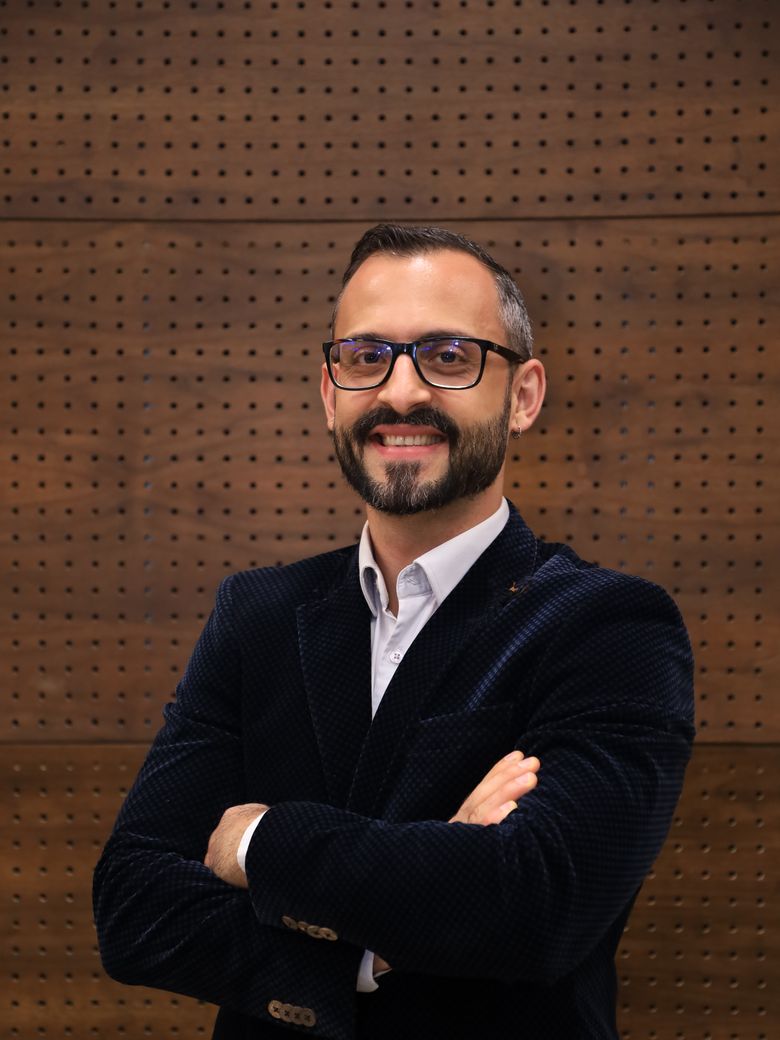 Caio Victor sorrindo, de óculos, blazer escuro e camisa branca, braços cruzados diante de uma parede de madeira.