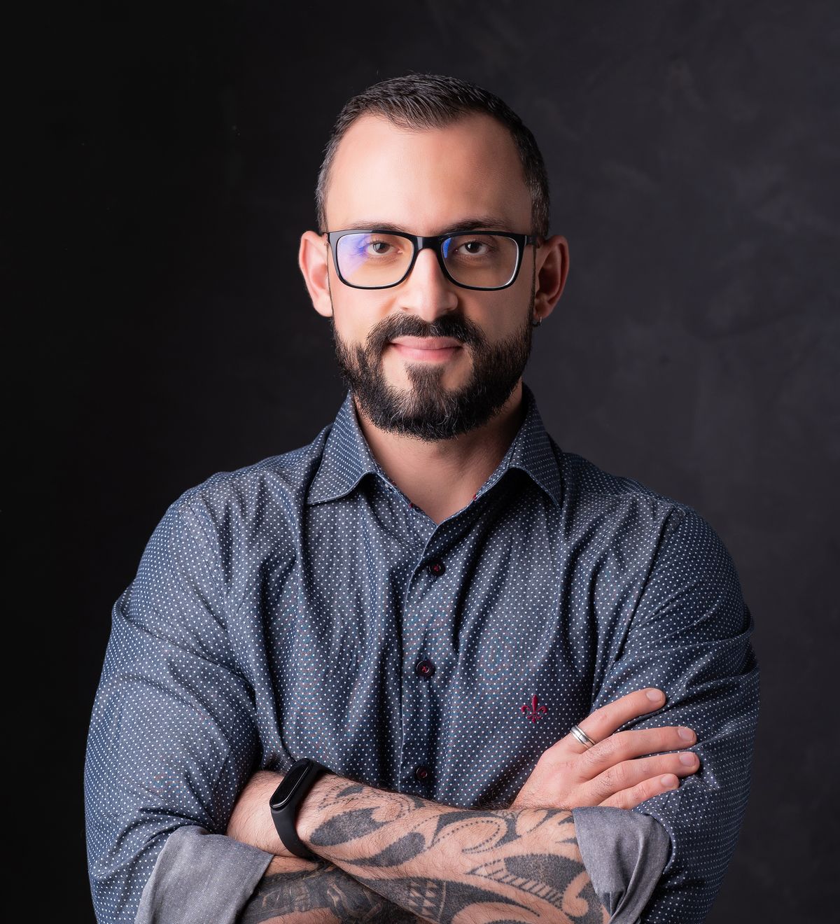 Caio Victor, de óculos e barba, camisa azul e braços cruzados, em estúdio de fundo escuro.